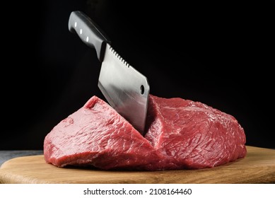 A piece of raw meat close-up with a chef's hatchet on a dark background. - Shutterstock ID 2108164460
