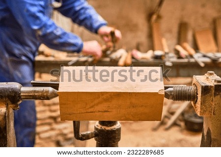 piece of wood prepared on a lathe to be turned by the carpenter who is in the background picking up the appropriate tool