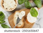 Pieces of bread with cream cheese and basil served on white marble table, flat lay