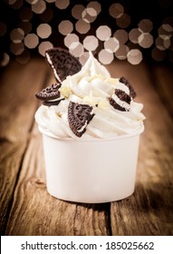 Pieces Of Broken Chocolate Oreos On A Twirl Of Rich Creamy Frozen Yoghurt In A Plastic Takeaway Tub On A Wooden Counter With A Bokeh Of Twinkling Lights