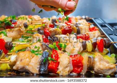 Pieces of chicken and vegetables on wooden skewers are fried to a rosy color. A female hand sprinkles a delicious hot dish with finely chopped leaves of cilantro, parsley and green onions.