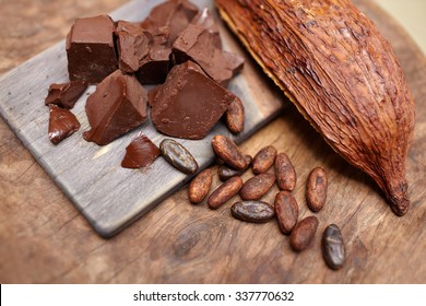 Pieces Of Chocolate With Roasted Cocoa Beans And Dry Cocoa Pod Fruit On A Wooden Table