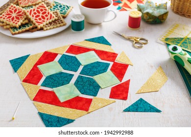 Pieces of fabric laid out in the shape of a patchwork block, a  heap of cookies with a pattern imitating a patchwork block, a cup of tea, sewing and quilting accessories