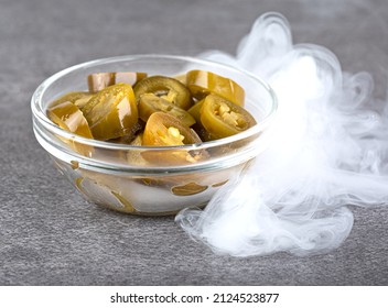 Pieces of pickled jalapeno pepper in a transparent bowl with smoke. Side view