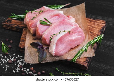 Pieces of raw pork steak with spices and herbs rosemary, thyme, basil, salt and pepper on dark black wooden background in rustic style