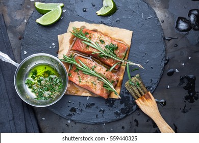 Pieces Of Raw Salmon Oiled Marinade With Spices, Lemon, Spices And Olive Oil With Brush On Slate Stone On A Dark Metallic Background. View From Above. Preparation For Cooking Fish Food. Salmon Steak.