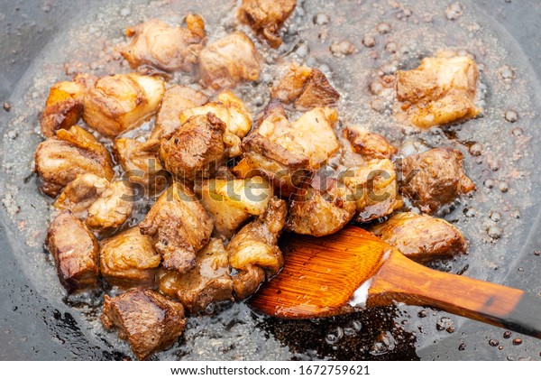 Pieces of red meat are fried in an old frying pan.\
Close up photo of meat, greaves, wooden kitchen spatula and\
splashes of fat.