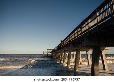 A pier extends over the ocean with waves crashing, under a clear blue sky at sunset, capturing a serene coastal scene.