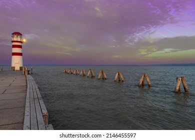 A pier leading to the lake and a red and white lighthouse on Lake Neusiedl in Podersdorf, Austria. In the background is a dramatic sky at sunrise.