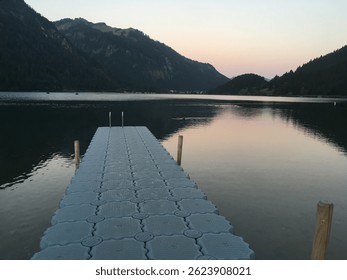 Pier or phonon as an entrance into fresh Alps lake in Austria during beautiful sunset colour sky. Mountains and sunset in reflecting in calm and crystal clear lake water. Spectacular Landscape for swi