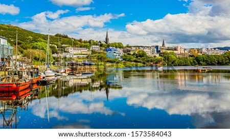 Pier at the port of Clifden at high tide, boats anchored with mirror reflection in the water, townscape against sky, sunny spring day with a blue sky and abundant white clouds in Clifden, Ireland