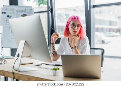 pierced businesswoman with pink hair holding pen and looking at laptop near computer monitor