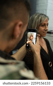 Piercing studio worker taking a cell phone photo of an older client with gold jewelry in the clips. Second youth concept. Vertical photo
