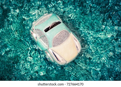 Piggy Bank Car With A Coin In It On Abstract Rustic Teal Blue Background Of Shattered Glass, View From Above. Money Savings For Emergency Events Or Vehicle Accident Insurance Conceptual Backdrop