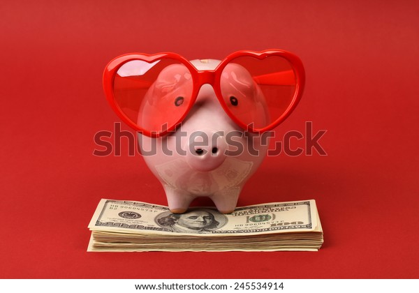 Piggy\
bank in love with red heart sunglasses standing on stack of money\
american hundred dollar bills on red\
background