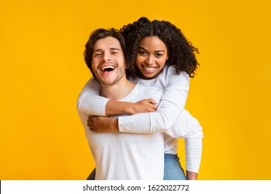 Piggyback ride. Affectionate guy carrying his black girlfriend on back, interracial couple bonding together over yellow background with free space - Powered by Shutterstock