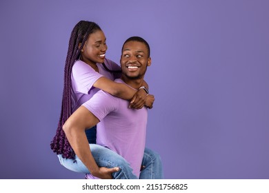 Piggyback Ride. Smiling black man carrying and riding his woman with long afro braids on back, enjoying time together, standing isolated on violet studio background, having fun, free copy space