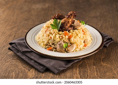 Pilaf With Chicken Drumstick In A Light Plate On A Napkin With Garlic And Fresh Herbs On A Dark Wooden Background. A Traditional Oriental Dish. Side View With Copy Space. Horizontal Orientation.