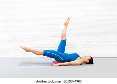 Pilates, abs and core workout with small fit ball. Fit caucasian woman in blue sportswear lying on back and practice single leg circles drill in loft fitness studio indoor.