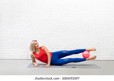 Pilates core and hips workout. Caucasian adult smiling woman lies on her side and raise her legs up holding a small fit ball between her feet, in loft studio indoor.
