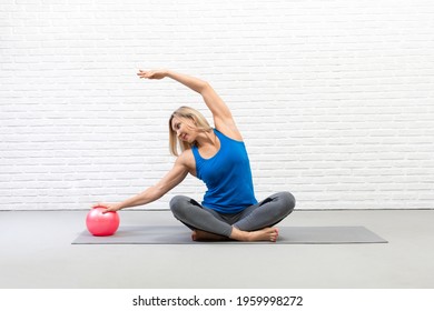 Pilates drill with mini-ball. Attractive adult caucasian woman in sportswear does sitting side bends using a small fit ball in loft studio.