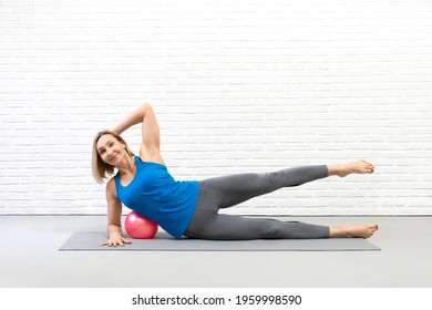 Pilates drill with small ball. Attractive fit caucasian smiling woman in sportswear does side leg lifts on forearm on the floor indoor, using a mini-ball.