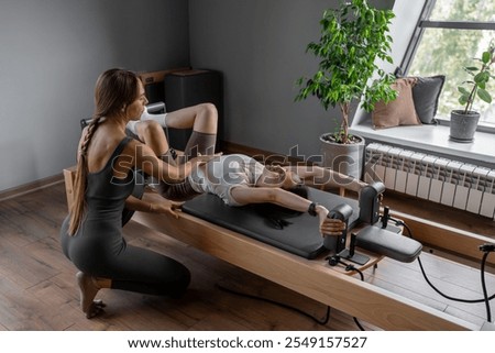 A Pilates Instructor helps a client with stretching exercises on reformer bed to improve flexibility and wellbeing 