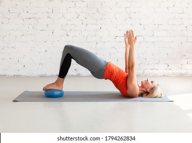Pilates with small fit ball. Caucasian woman practice gluteal bridge balance on a mat, in loft white studio indoor, selective focus. Workout, fitness, coach, healthy lifestyle concept.