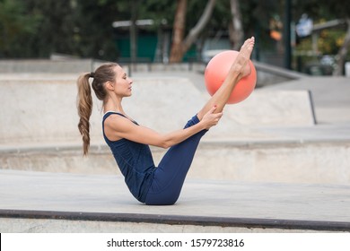 Pilates teacher with a slim athletic body and beautiful face, wearing dark blue clothes, holding a V pose with a small orange Pilates ball between her legs. Side view with Urban skate park background. - Powered by Shutterstock