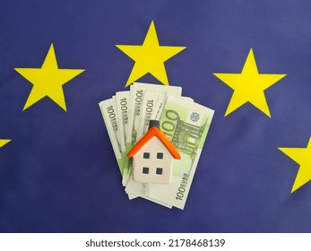 Pile Of 100 Euro Banknotes And Small Toy House On European Flag. Representation Of Housing Costs In European Union EU And Mortgage Costs Concept