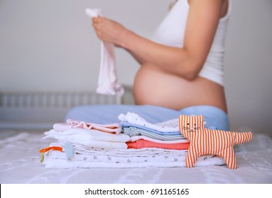 Pile Of Baby Clothes, Necessities And Pregnant Woman On Bed In Home Interior Of Bedroom. Pregnant Woman Is Getting Ready For The Maternity Hospital, Packing Baby Stuff. Pregnancy, Birth Concept.