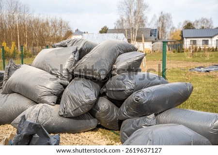 A pile of black plastic bags rests on a gravel surface, enclosed by a green wire fence. Nearby are residential houses, a greenhouse, and leafless trees.