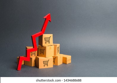 A Pile Of Cardboard Boxes With Drawing Of Shopping Carts And A Red Up Arrow. The Growth Rate Of Production Of Goods And Products, Increasing Economic Indicators. Increasing Consumer Demand, Exports