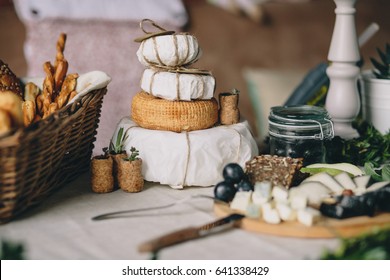 A pile of cheese heads in a paper, next a sliced cheese, a basket of bread and grapes, wine and plant plugs