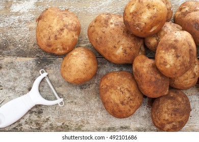 Pile Of Farm Fresh Potatoes Still With Adhering Dirt And A Manual Peeler Lying On A Rustic Background Ready To Prepare For Dinner