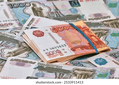 A pile of five thousand ruble notes, tied up with a green rubber band, lies on top of a large number of thousand ruble notes. The growth of citizens' wealth. Increase in savings income.