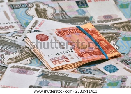 A pile of five thousand ruble notes, tied up with a green rubber band, lies on top of a large number of thousand ruble notes. The growth of citizens' wealth. Increase in savings income.