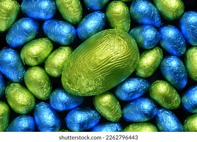 Pile or group of multi colored and different sizes of colourful foil wrapped chocolate easter eggs in blue, yellow and lime green with a large green egg in the middle. 