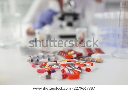 Pile of pills scattered on white table against woman doing research with microscope. Medicine and scientific research in laboratory
