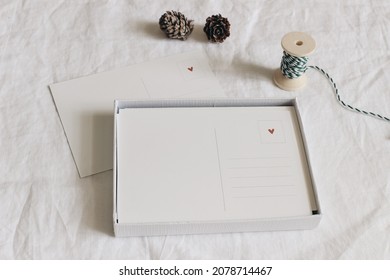 Pile Of Postcards With Red Heart In Paper Box. Blank Greeting Cards Mockups. Pine Cones And Decorative Gift Rope. White Blurred Linen Table Cloth Background. Christmas Stationery. Winter Top View