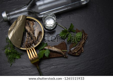 A pile of pot vodka with sprats, a sandwich with black bread, a fork and a bottle on a dark background. View from the top 