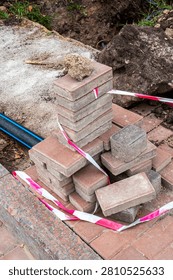 A pile of red and gray interlocking paving stones are stacked on a construction site, cordoned off with red and white warning tape