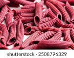 A pile of red uncooked plant-based beet penne pasta on a white ceramic plate close up. Organic gluten free ingredient