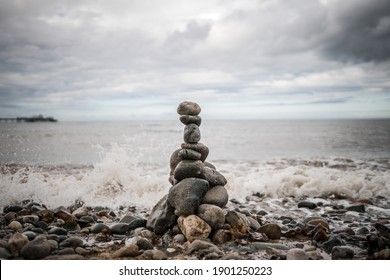 Pile of stones stacked up balancing sea shore ocean lapping up pebbles tower grey granite with dramatic storm clouds gather above waves crashing on the shore at seaside as tide comes in holiday resort