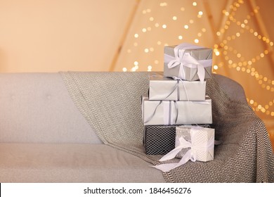 Pile Of Stylishly Wrapped Gift Boxes Placed On Couch With Blanket In Cozy Room Against Wall With Fairy Lights On Christmas Day