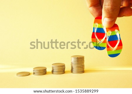 Piles of coins are laid out in increasing number of coins in a stack on a yellow background and a hand holding beach slippers. Concept - the accumulation of money on vacation, vacation budget.