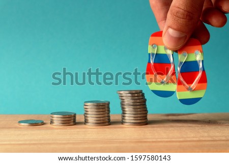 Piles of coins are laid out in increasing number of coins in a stack on a blue background and a hand holding beach slippers. Concept - the accumulation of money on vacation, vacation budget.