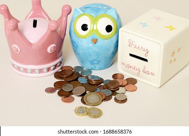 Piles Of UK Currency Coins, Including Pennies And Pounds, Emptied Out In Front Of Pretty Pink, Blue And Cream Children's Money Boxes.  Pretty Piggy Banks For Children's Savings Pocket Money