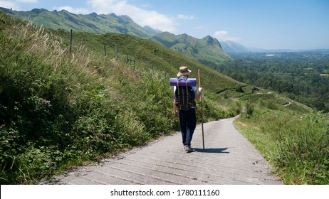 Pilgrim with hat, backpack and stick walking a road between a mountain and a village under the blue sky
