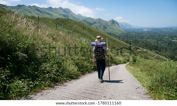 Pilgrim with hat, backpack\
and stick walking a road between a mountain and a village under the\
blue sky